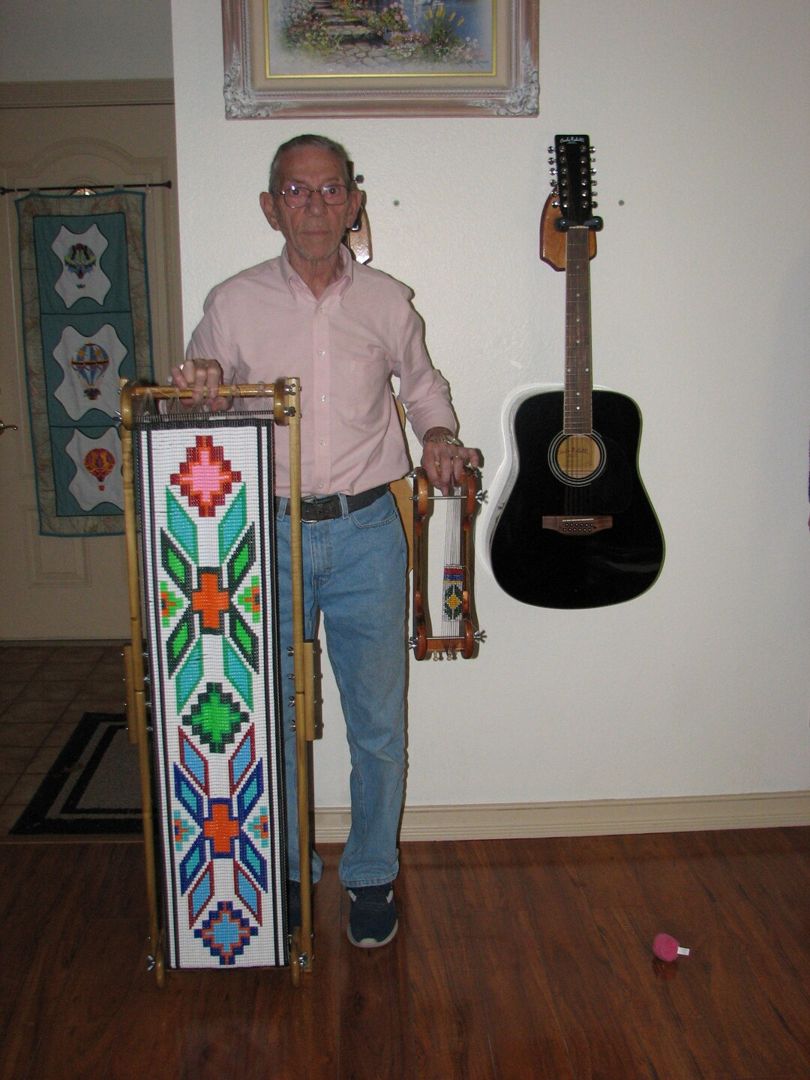 A man standing beside a tall loom that holds a long finished beadwork panel